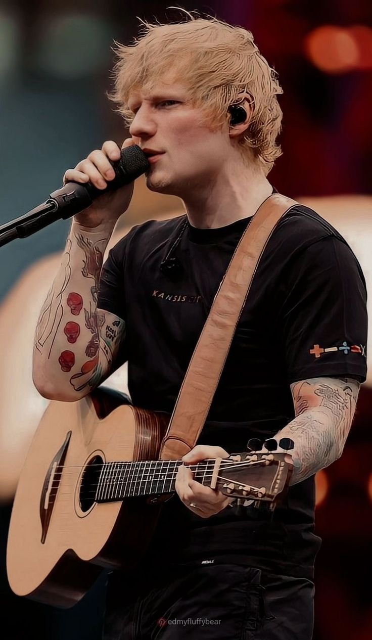 Ed Sheeran performing live on stage with a guitar, singing into the microphone under vibrant stage lights.